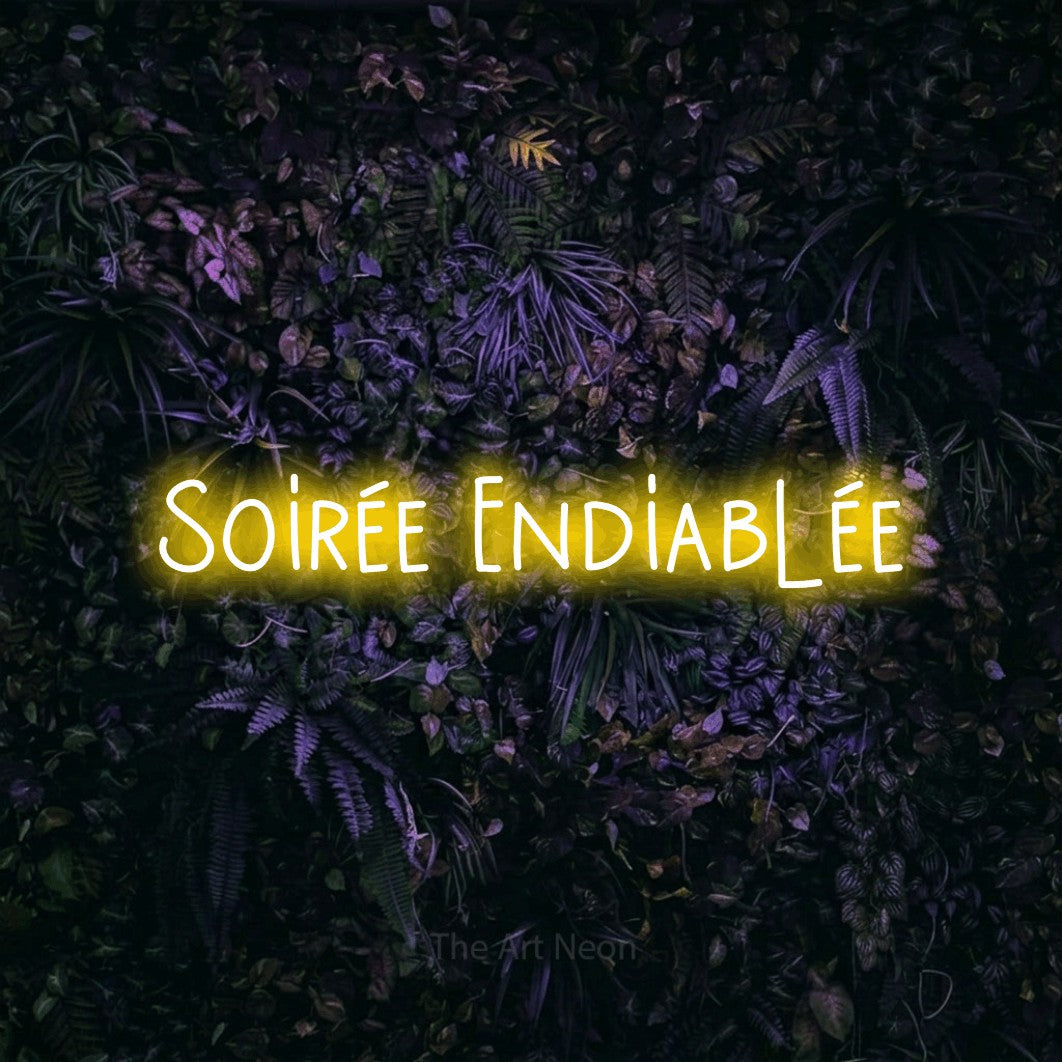“Soirée endiablée” LED neon sign in yellow, with narrow hand-drawn lettering, shown against a dark wall with purple flowers.