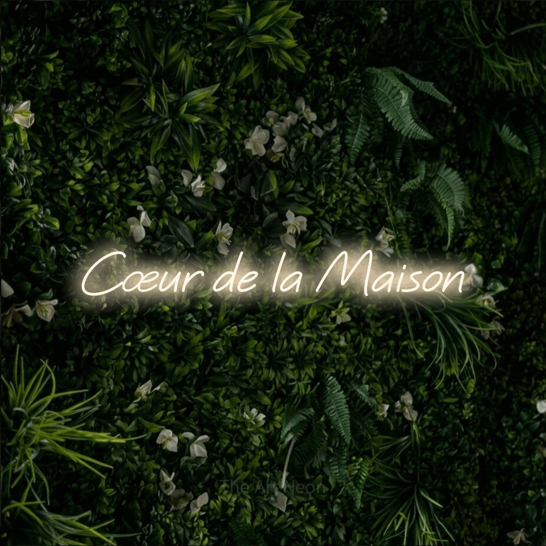 “Cœur de la maison” LED neon sign in warm white, with slanted cursive lettering, shown against a dense green plant wall.