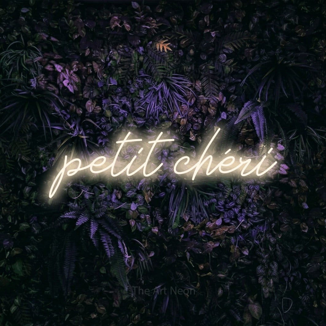 “Petit chéri” LED neon sign in warm white, with cursive lettering, shown against a dark wall with purple flowers.