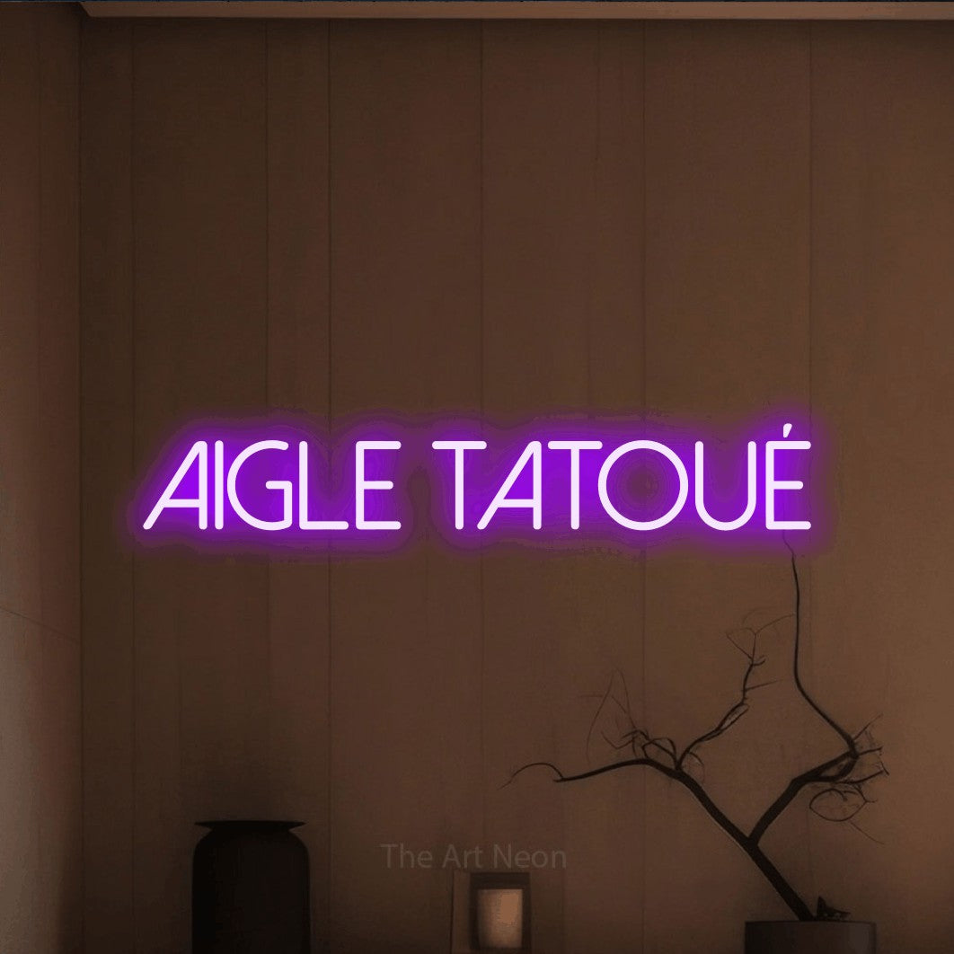 “Aigle tatoué” LED neon sign in purple, with thin sans-serif lettering, shown against a simple earth-toned interior.