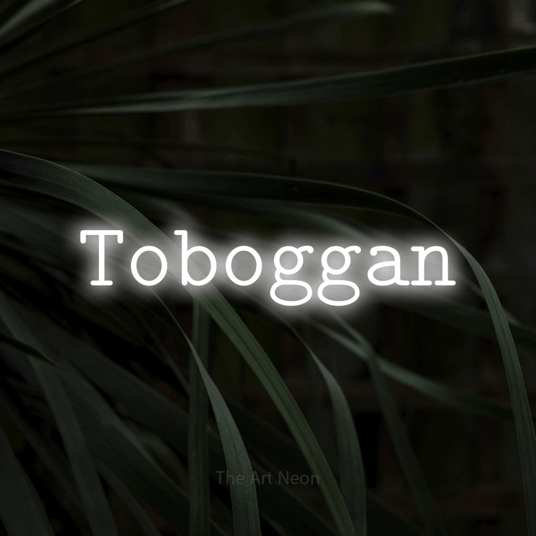 “Toboggan” LED neon sign in cool white, with typewriter-style lettering, shown against a close-up of dark foliage.