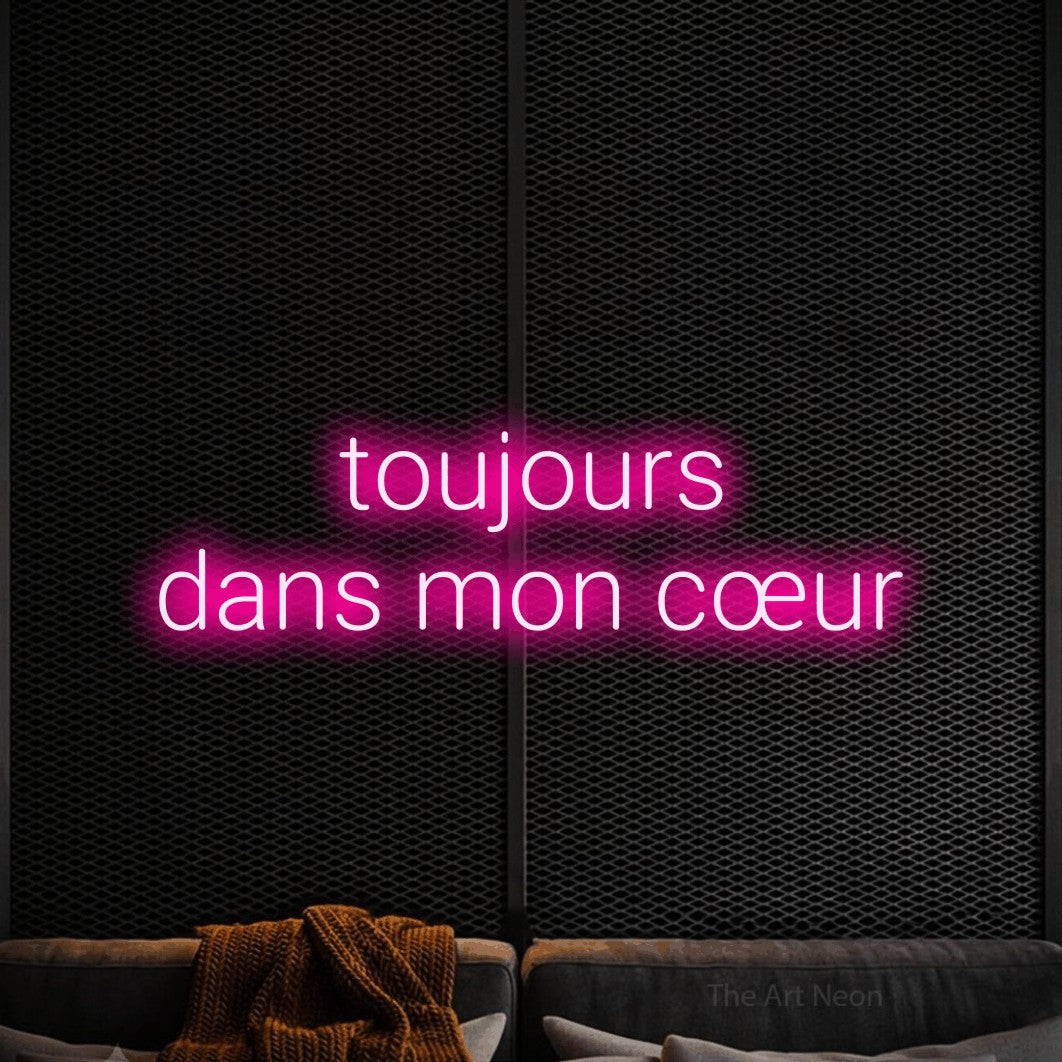 “Toujours dans mon cœur” LED neon sign in hot pink, with rounded sans-serif lettering across 2 lines, shown against a modern dark-toned seating area.