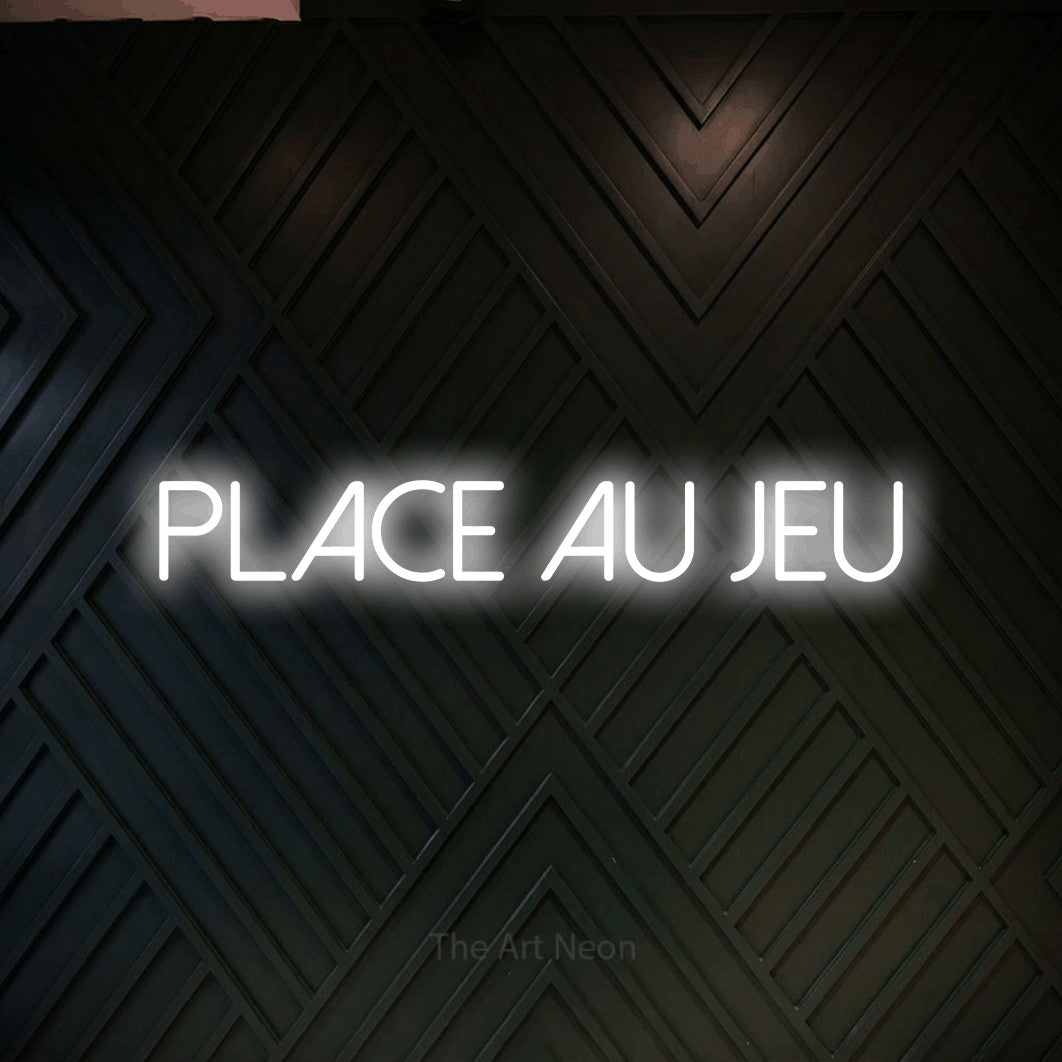 “Place au jeu” LED neon sign in cool white, with thin sans-serif lettering, shown against a black chevron-patterned wood wall.