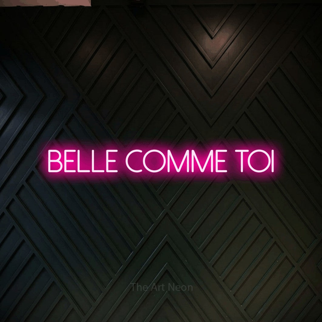 Lettrage en néon LED « Belle comme toi » en rose vif, en lettres fines sans empattement, présenté sur un mur en bois noir à motif chevron, pour un salon de beauté.
