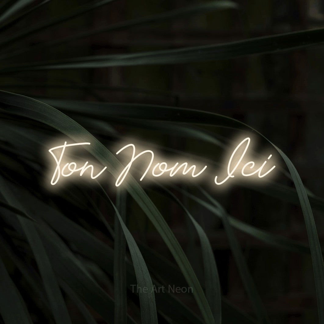 “Ton nom ici” LED neon sign in warm white, with cursive lettering, shown against a close-up of dark foliage.