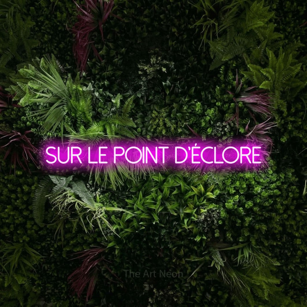 “Sur le point d'éclore” LED neon sign in pink, with thin sans-serif lettering, shown against a plant wall with pink flowers.