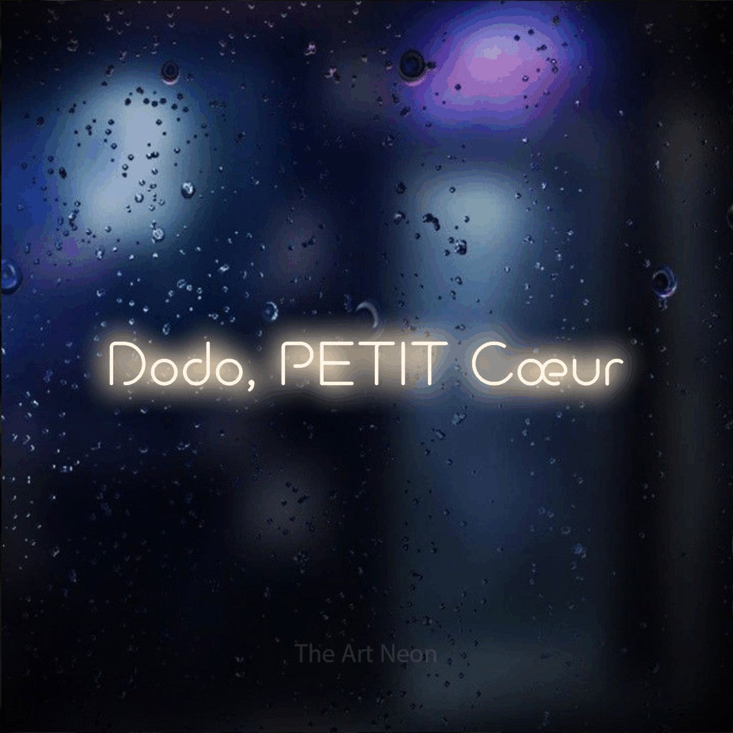 “Dodo, petit cœur” LED neon sign in warm white, with stylised geometric lettering, shown against glass with blue and purple reflections.