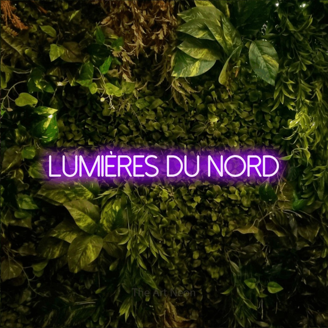 “Lumières du nord” LED neon sign in purple, with thin sans-serif lettering, shown against bright green and yellow foliage.