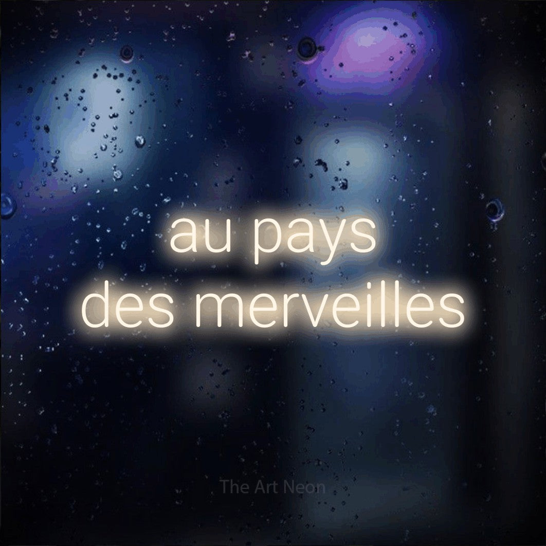 “Au pays des merveilles” LED neon sign in warm white, with rounded sans-serif lettering across 2 lines, shown against glass with blue and purple reflections.