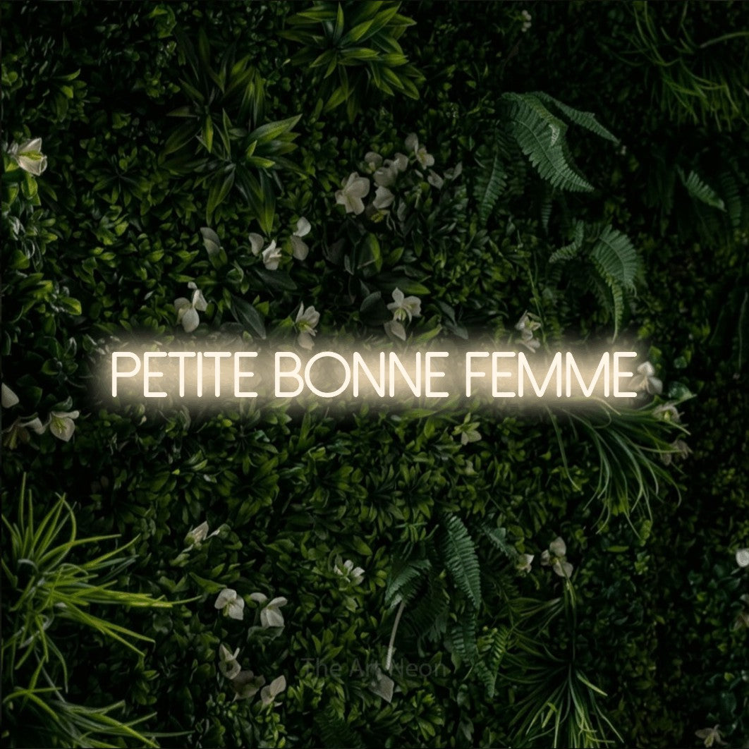 “Petite bonne femme” LED neon sign in warm white, with thin sans-serif lettering, shown against a dense green plant wall.