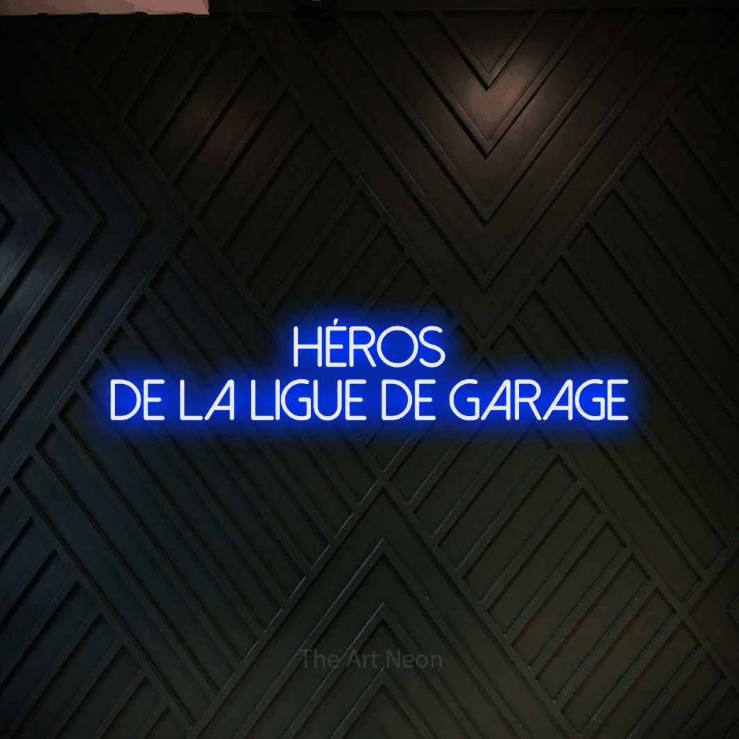 “Héros de la ligue de garage” LED neon sign in blue, with thin sans-serif lettering across 2 lines, shown against a black chevron-patterned wood wall.