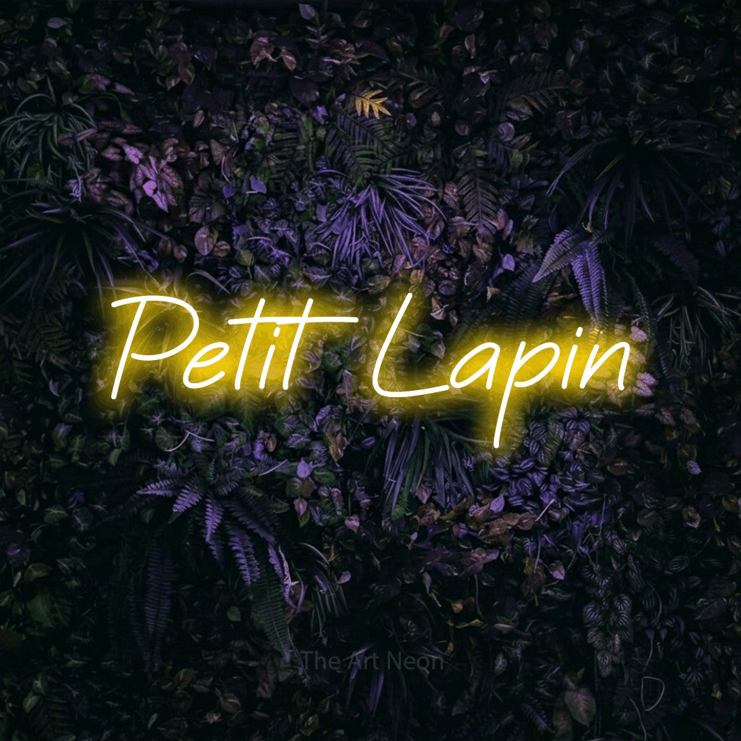 “Petit lapin” LED neon sign in yellow, with slanted cursive lettering, shown against a dark wall with purple flowers.