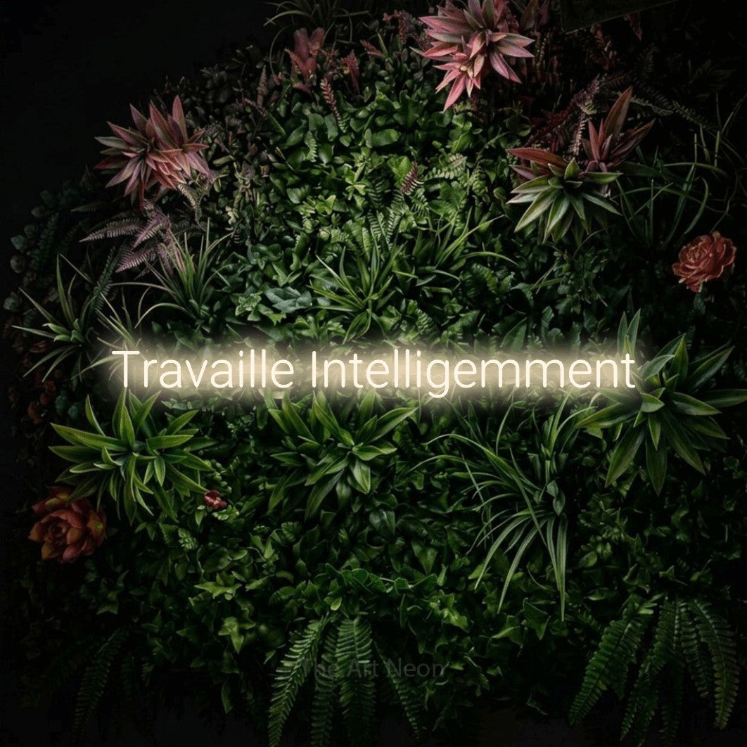 “Travaille intelligemment” LED neon sign in warm white, with rounded sans-serif lettering, shown against a flowering plant wall.