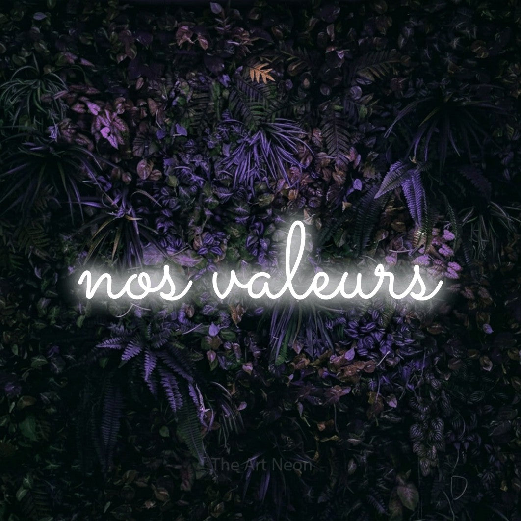 “Nos valeurs” LED neon sign in cool white, with looped cursive lettering, shown against a dark wall with purple flowers.