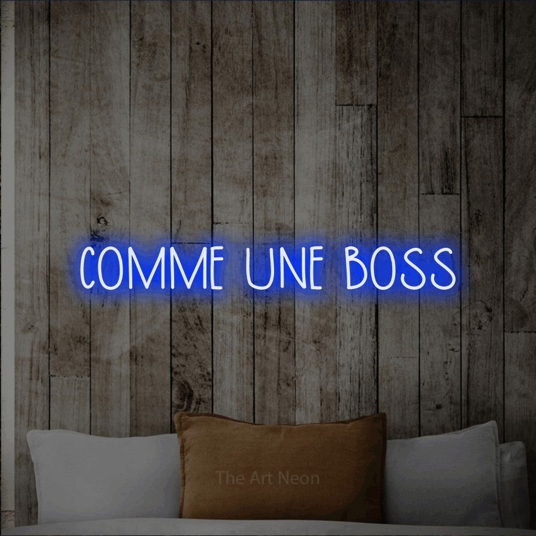 “Comme une boss” LED neon sign in blue, with narrow hand-drawn lettering, shown against a light rustic wood wall.