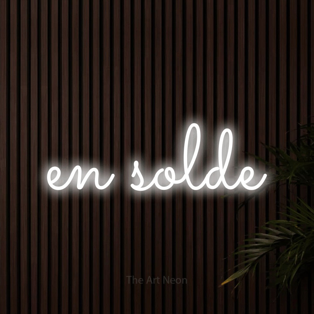 “En solde” LED neon sign in cool white, with looped cursive lettering, shown against dark wooden wall slats.