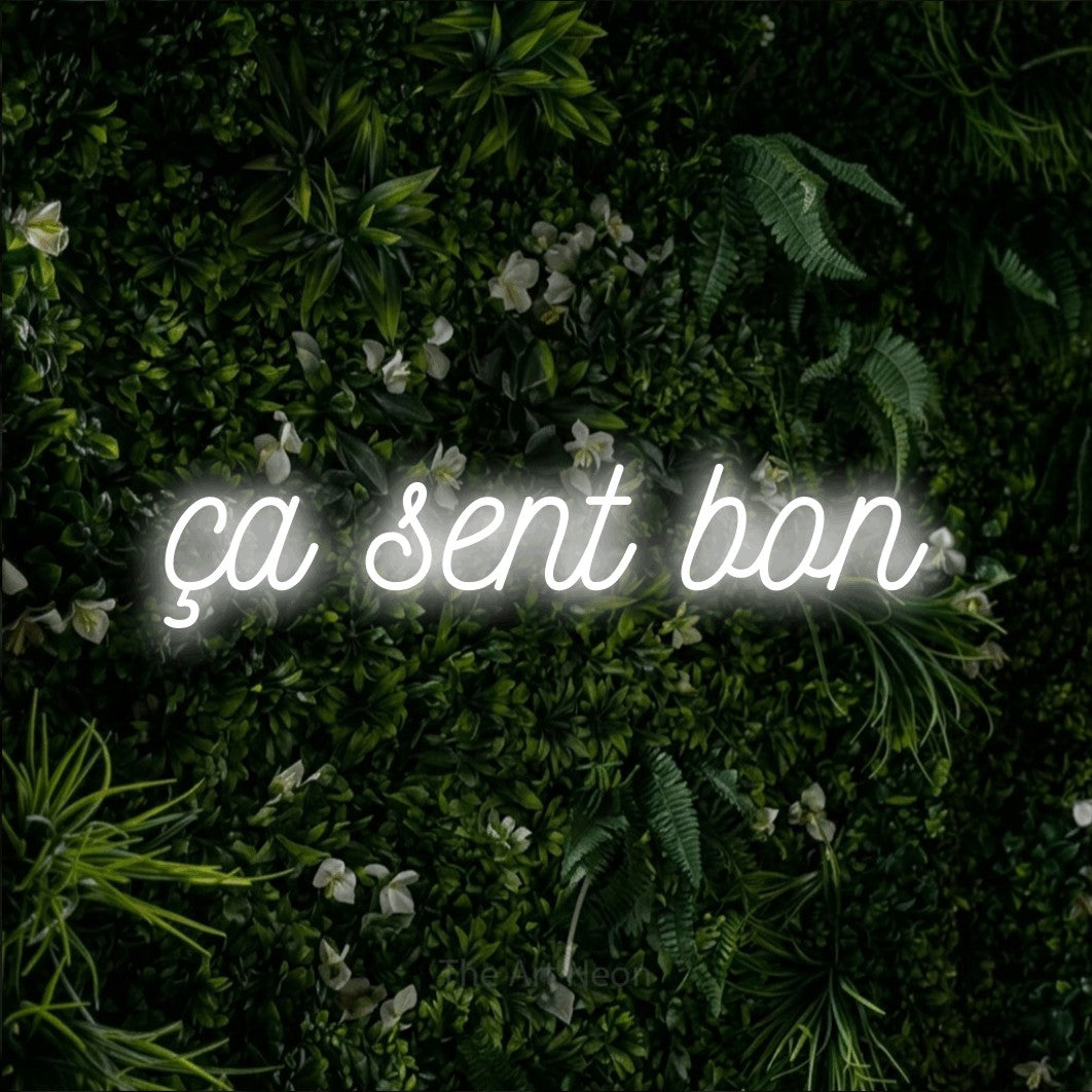 “Ça sent bon” LED neon sign in cool white, with ornate cursive lettering, shown against a dense green plant wall.