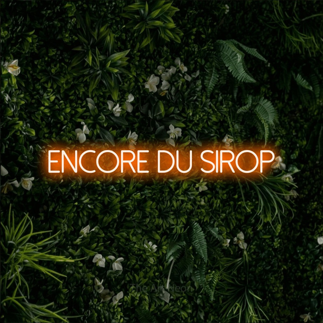 “Encore du sirop” LED neon sign in orange, with thin sans-serif lettering, shown against a dense green plant wall.