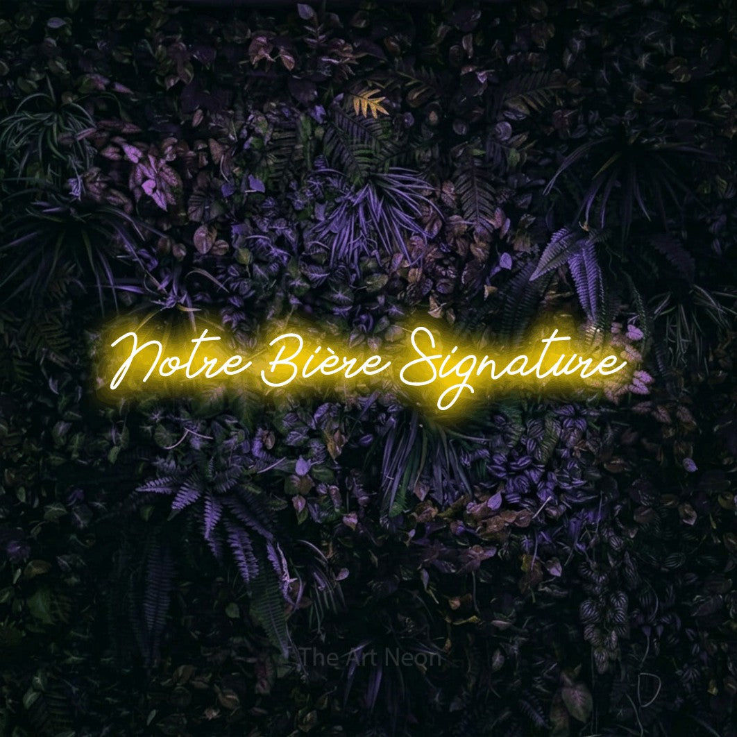 “Notre bière signature” LED neon sign in yellow, with cursive lettering, shown against a dark wall with purple flowers.