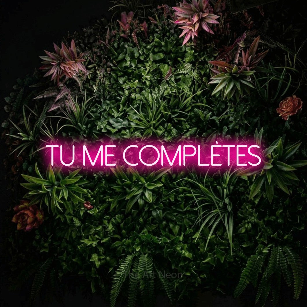 “Tu me complètes” LED neon sign in hot pink, with thin sans-serif lettering, shown against a flowering plant wall.