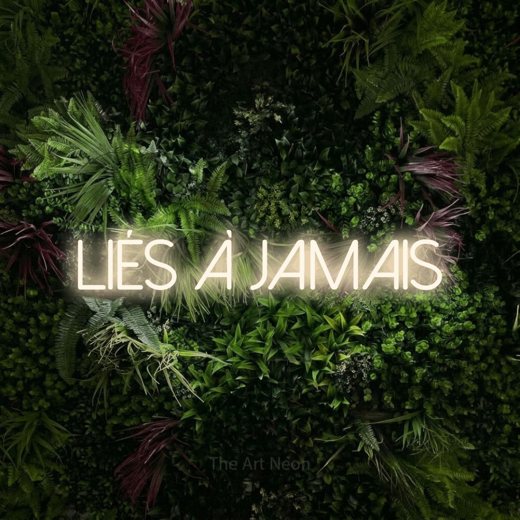 “Liés à jamais” LED neon sign in warm white, with thin sans-serif lettering, shown against a plant wall with pink flowers.