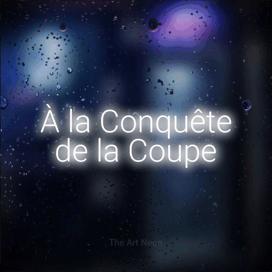 “À la conquête de la coupe” LED neon sign in cool white, with rounded sans-serif lettering across 2 lines, shown against glass with blue and purple reflections.
