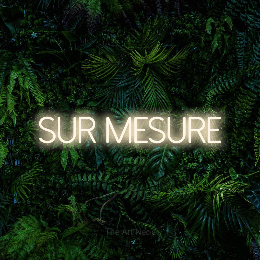 “Sur mesure” LED neon sign in warm white, with thin sans-serif lettering, shown against dense tropical foliage.