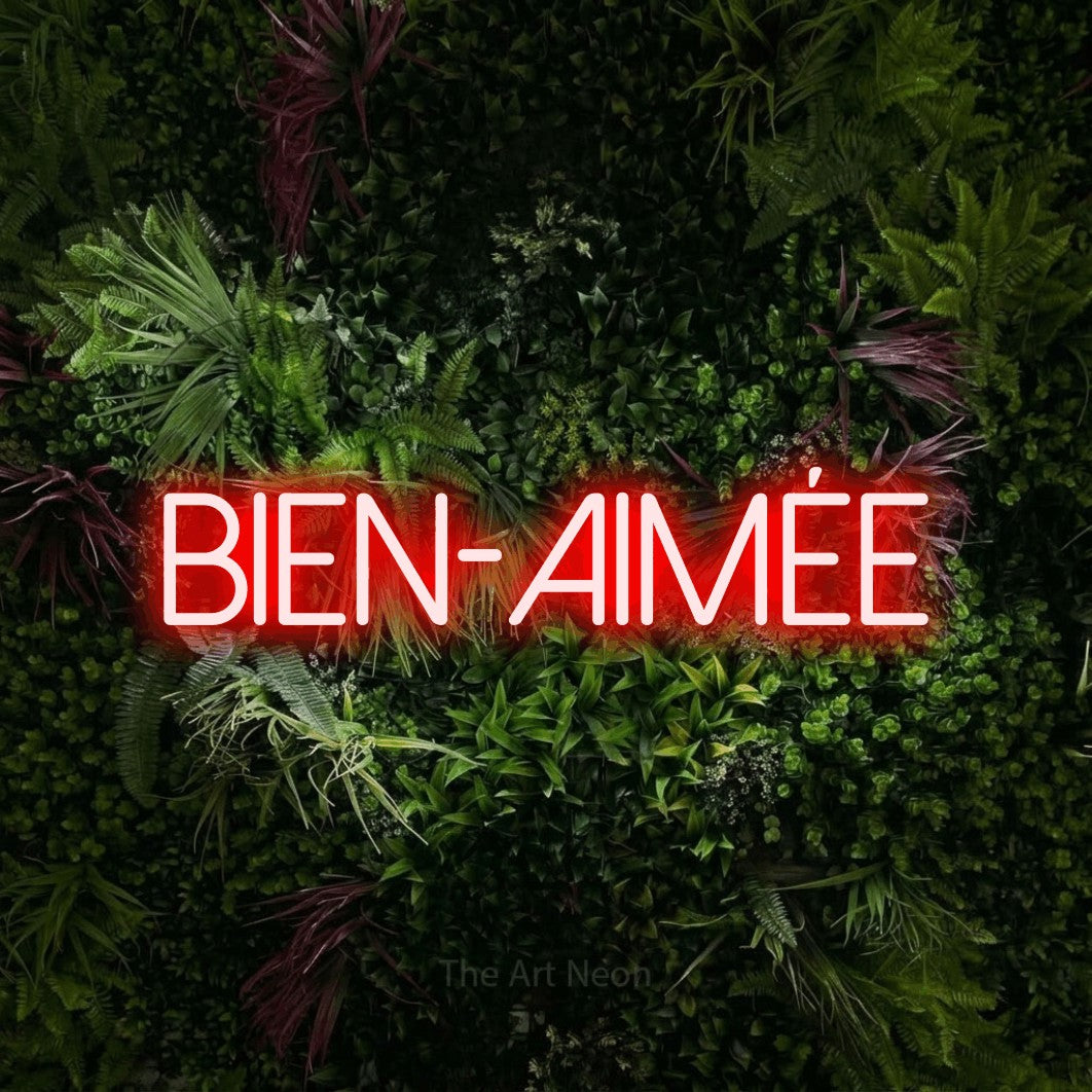 Aperçu d’une enseigne néon LED « Bien-aimée » en rouge, en lettres fines sans empattement, photographiée sur un mur végétal aux fleurs roses, pour un décor romantique.