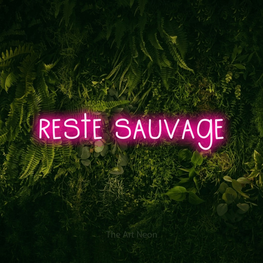 “Reste sauvage” LED neon sign in hot pink, with narrow hand-drawn lettering, shown against a dark moss and forest backdrop.