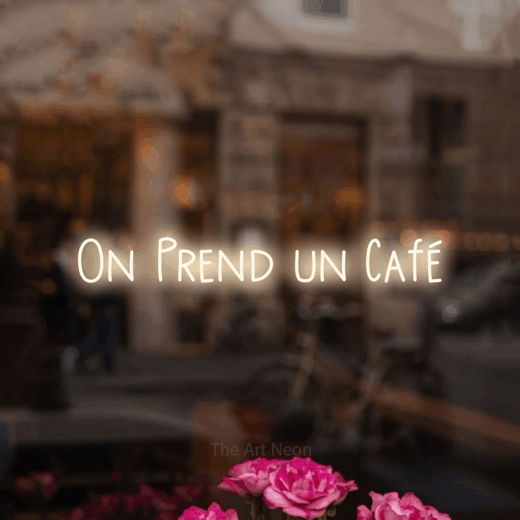 “On prend un café” LED neon sign in warm white, with narrow hand-drawn lettering, shown against a blurred café-bar interior.