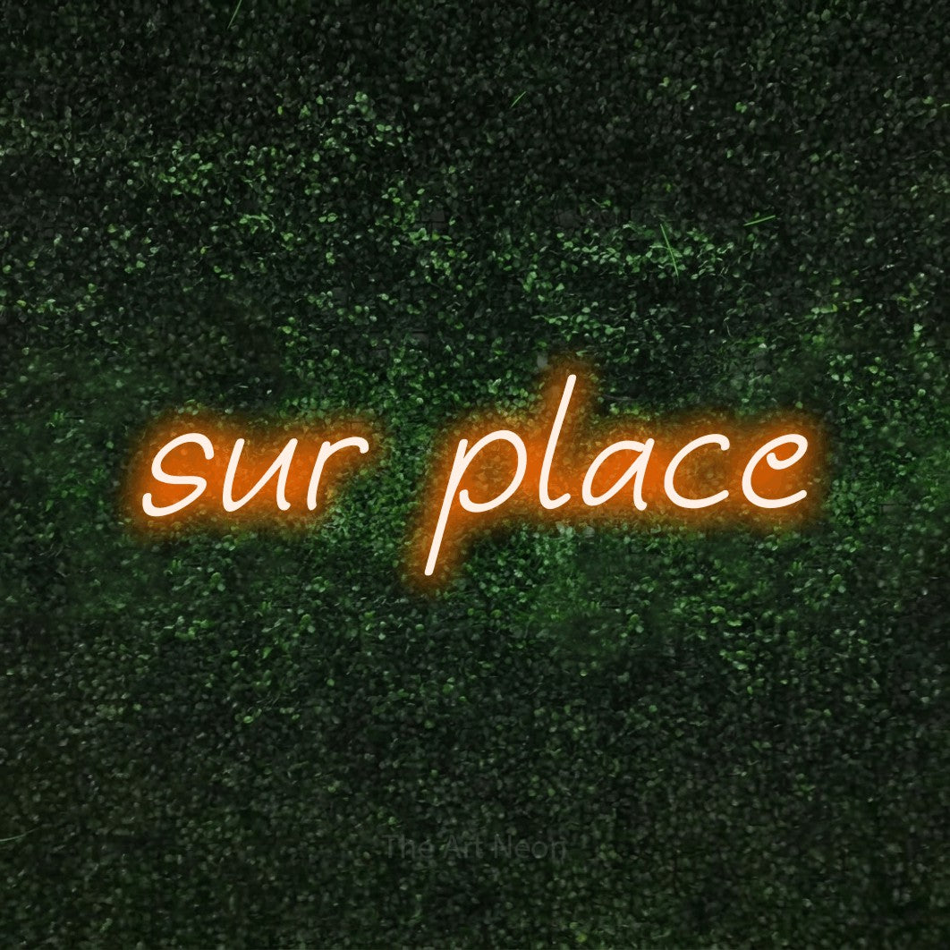 “Sur place” LED neon sign in orange, with slanted handwritten lettering, shown against a textured green hedge.