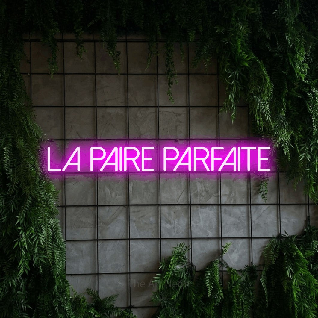 Aperçu d’une enseigne néon LED « La paire parfaite » en rose, en lettres fines sans empattement, photographiée sur une grille de béton couverte de lierre, pour une réception de mariage.
