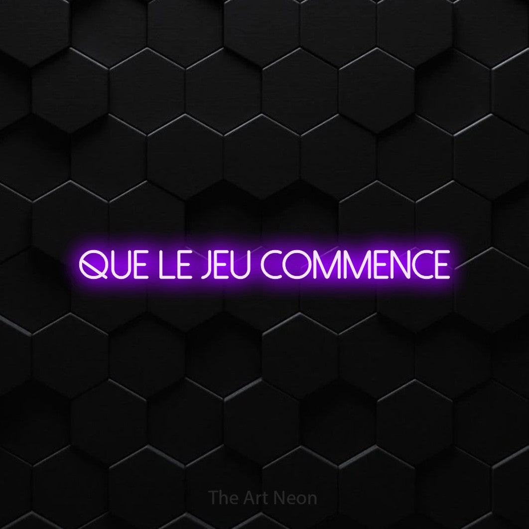 “Que le jeu commence” LED neon sign in purple, with thin sans-serif lettering, shown against black hexagonal wall tiles.
