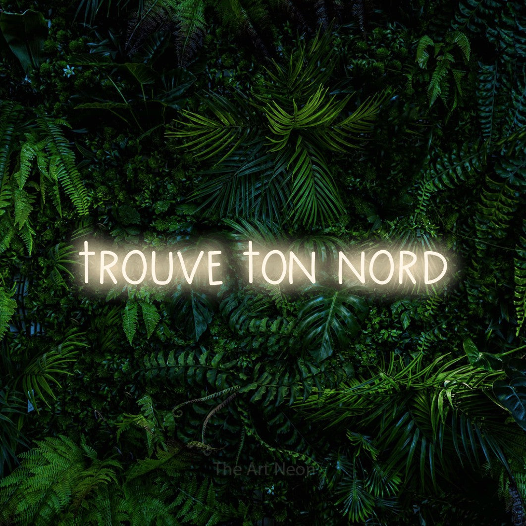 “Trouve ton nord” LED neon sign in warm white, with narrow hand-drawn lettering, shown against dense tropical foliage.