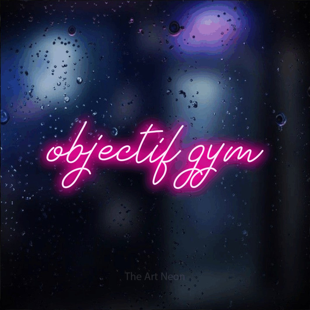 Néon LED « Objectif gym » en rose vif, en lettrage cursif, mis en scène sur une vitre aux reflets bleus et violets, pour un espace d’entraînement.