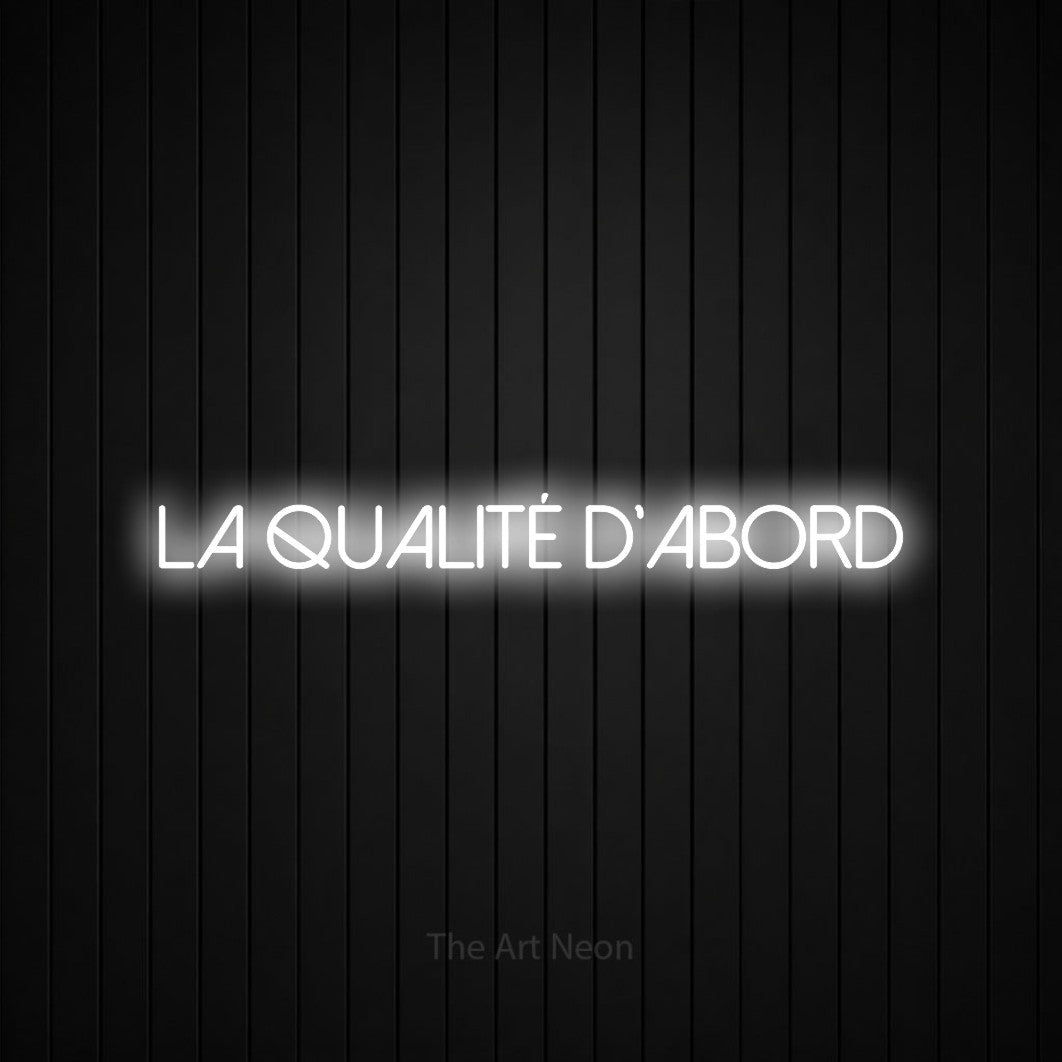 “La qualité d'abord” LED neon sign in cool white, with thin sans-serif lettering, shown against black vertical wall slats.