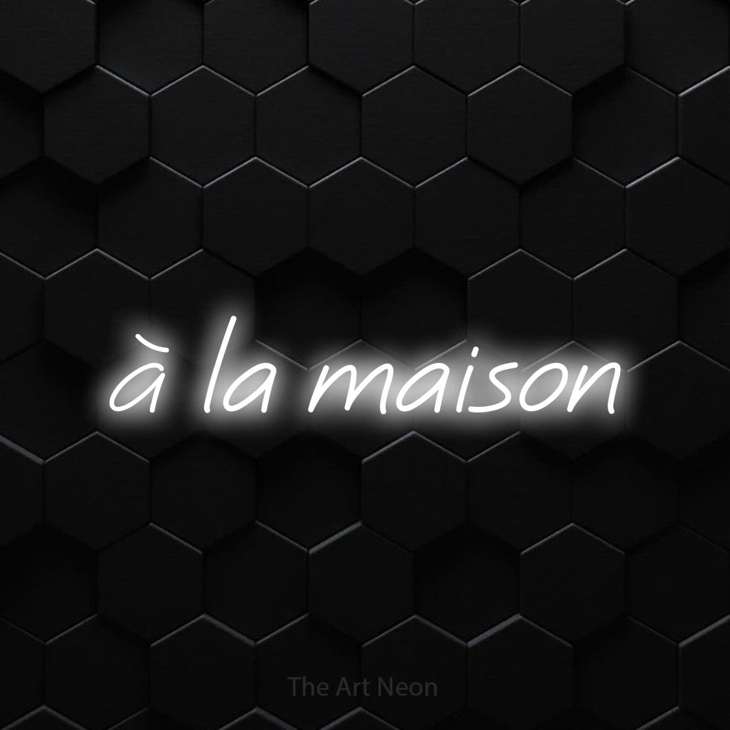 “À la maison” LED neon sign in cool white, with slanted cursive lettering, shown against black hexagonal wall tiles.