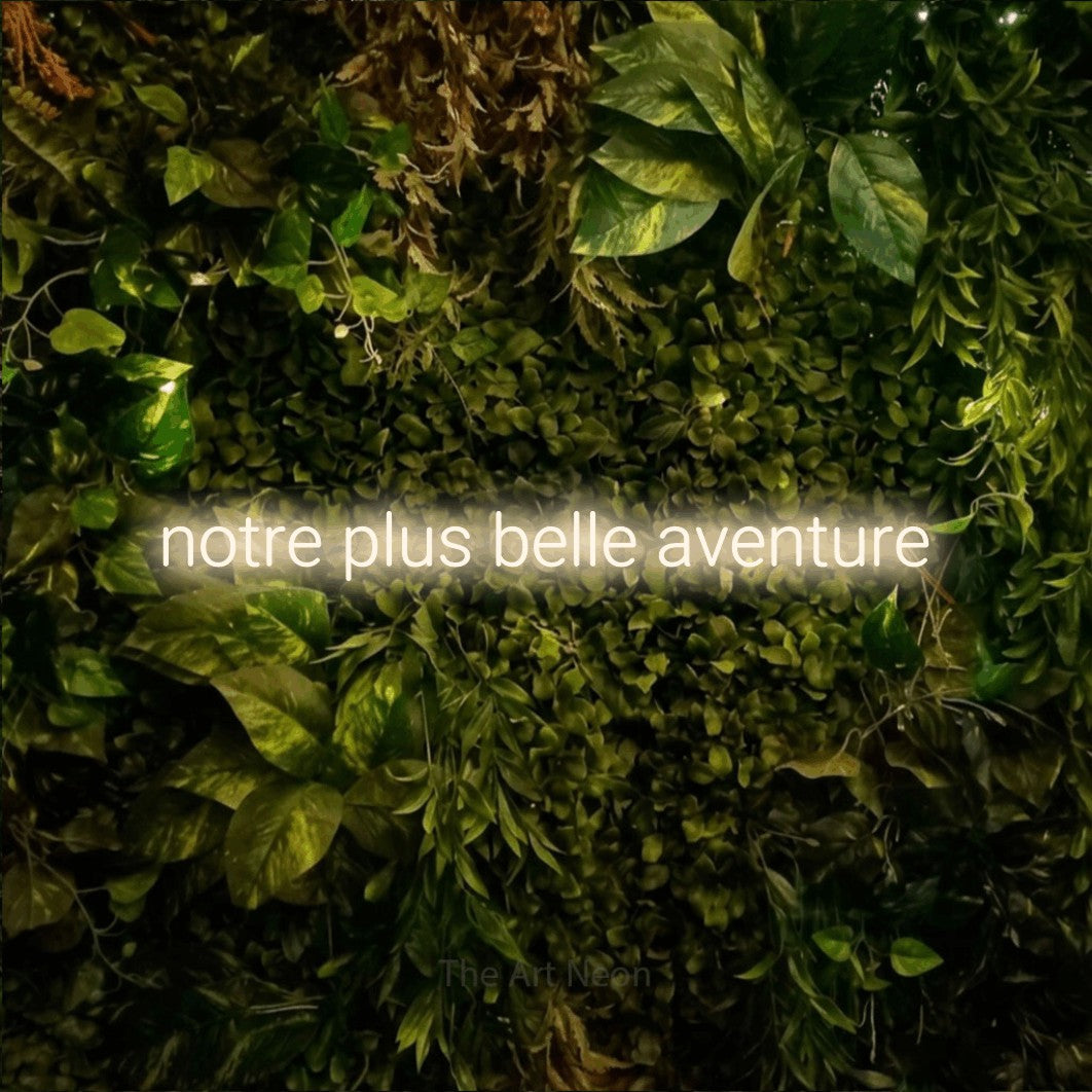 Lettrage en néon LED « Notre plus belle aventure » en blanc chaud, en lettres sans empattement arrondies, présenté sur un feuillage vert et jaune vif, pour une fête prénatale.