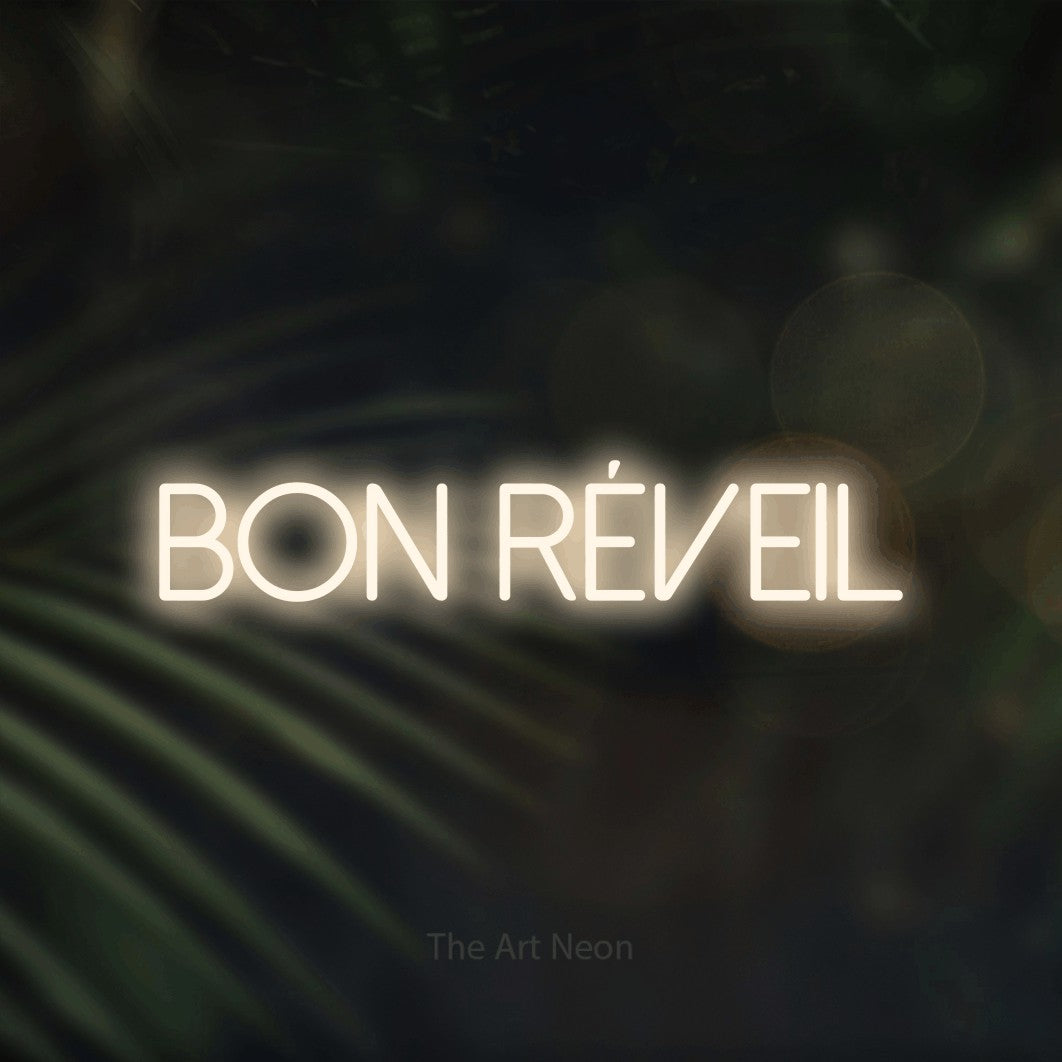 “Bon réveil” LED neon sign in warm white, with thin sans-serif lettering, shown against a blurred palm and bokeh-light backdrop.