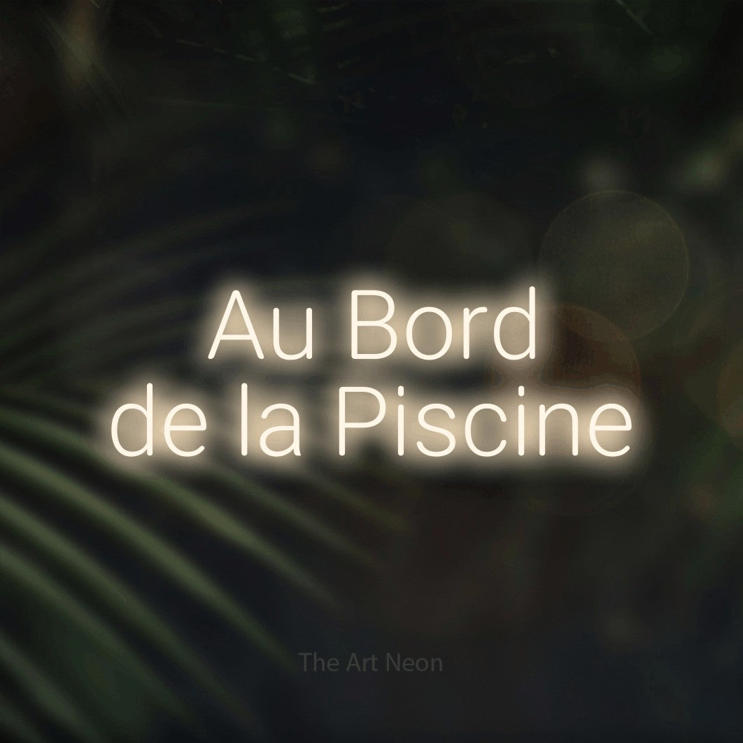 “Au bord de la piscine” LED neon sign in warm white, with rounded sans-serif lettering across 2 lines, shown against a blurred palm and bokeh-light backdrop.
