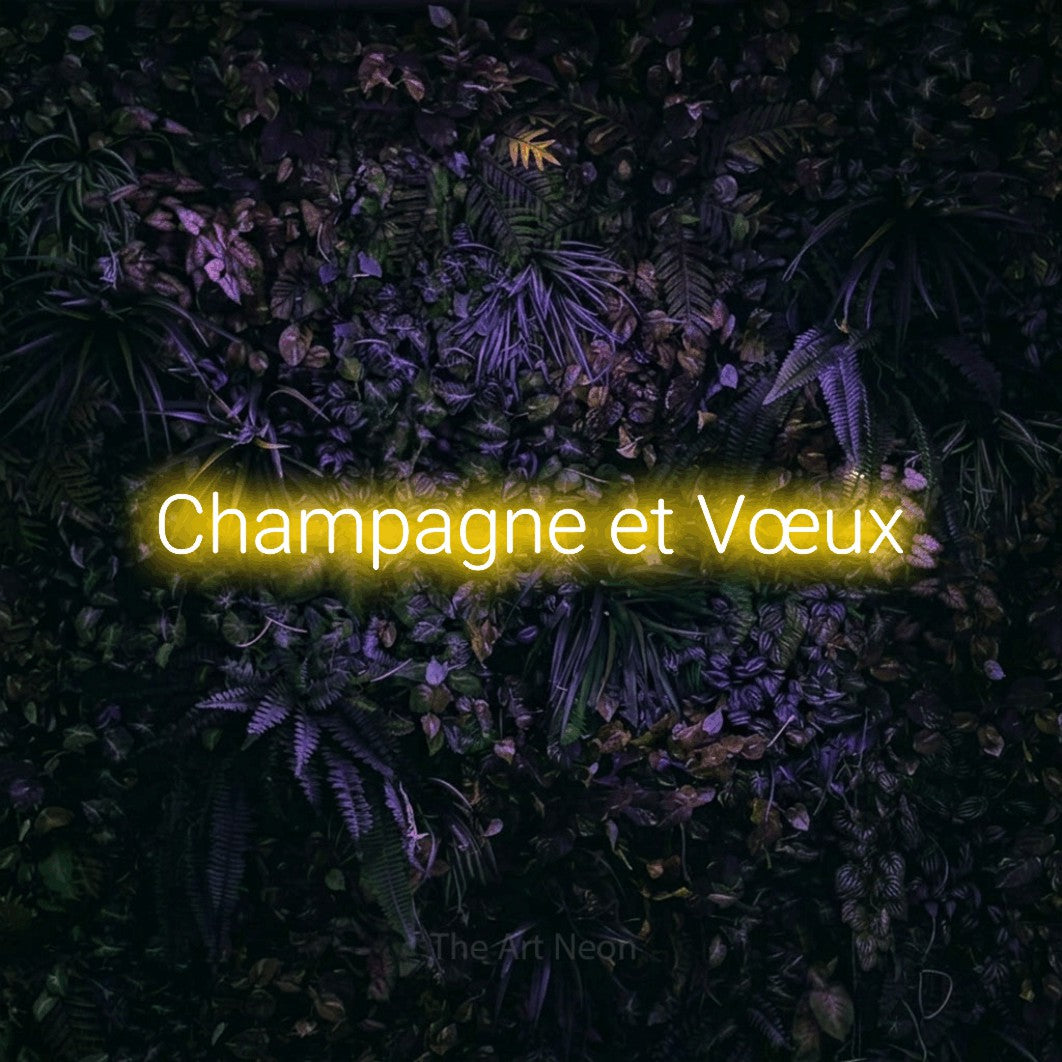 “Champagne et vœux” LED neon sign in yellow, with rounded sans-serif lettering, shown against a dark wall with purple flowers.