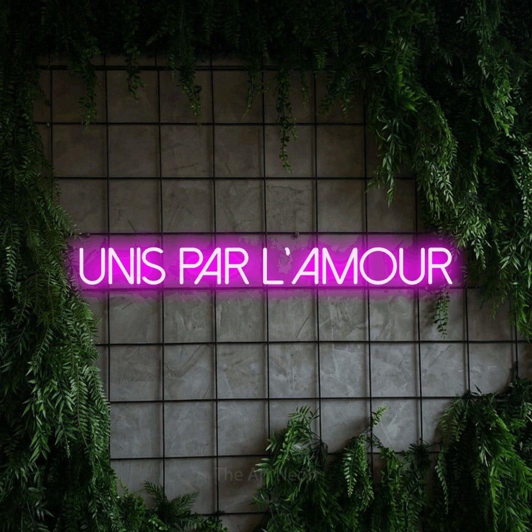 “Unis par l'amour” LED neon sign in pink, with thin sans-serif lettering, shown against an ivy-covered concrete grid.