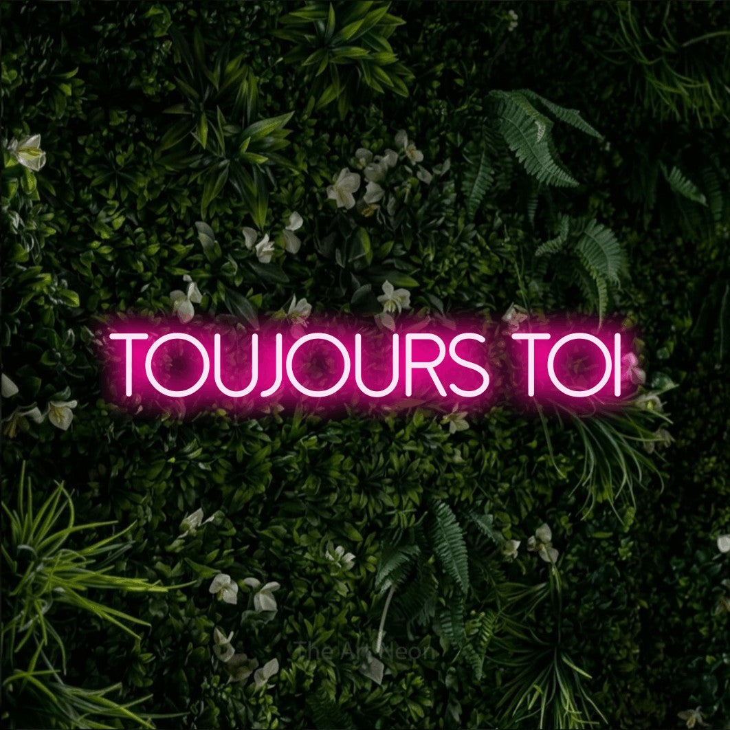 Néon LED « Toujours toi » en rose vif, en lettres fines sans empattement, mis en scène sur un mur végétal vert et dense, pour un décor romantique.