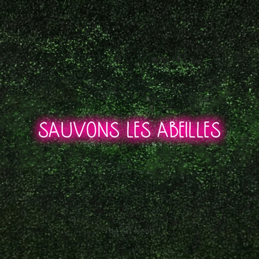 “Sauvons les abeilles” LED neon sign in hot pink, with narrow hand-drawn lettering, shown against a textured green hedge.