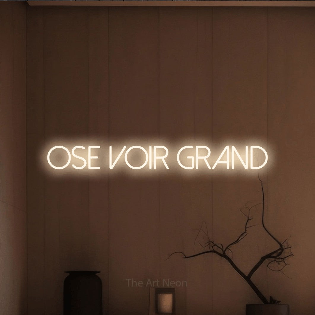 “Ose voir grand” LED neon sign in warm white, with thin sans-serif lettering, shown against a simple earth-toned interior.
