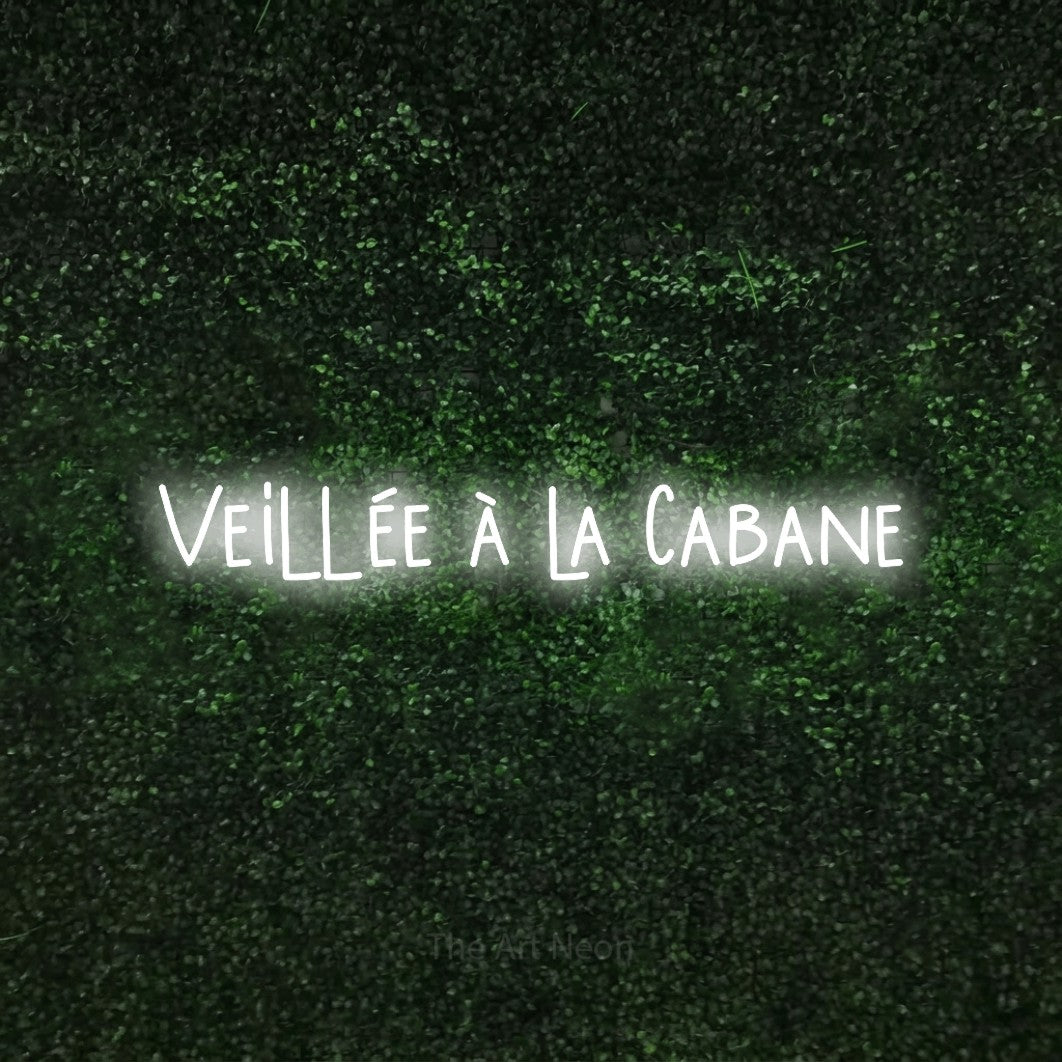 “Veillée à la cabane” LED neon sign in cool white, with narrow hand-drawn lettering, shown against a textured green hedge.