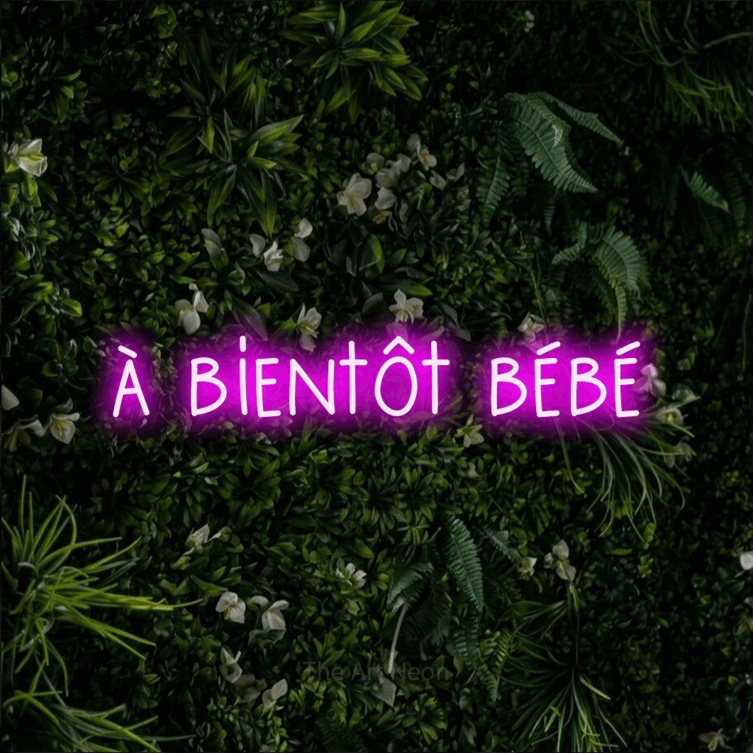 “À bientôt bébé” LED neon sign in pink, with narrow hand-drawn lettering, shown against a dense green plant wall.