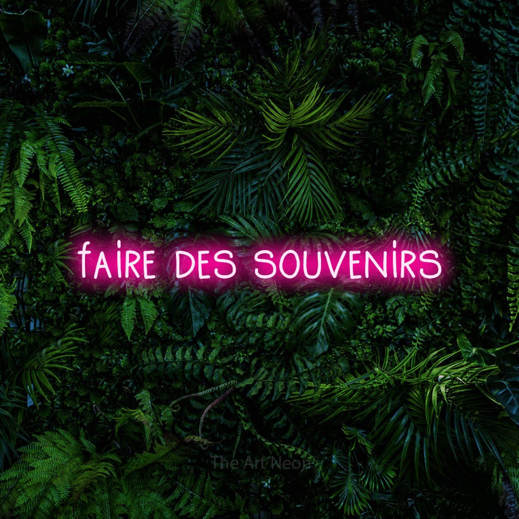 “Faire des souvenirs” LED neon sign in hot pink, with narrow hand-drawn lettering, shown against dense tropical foliage.