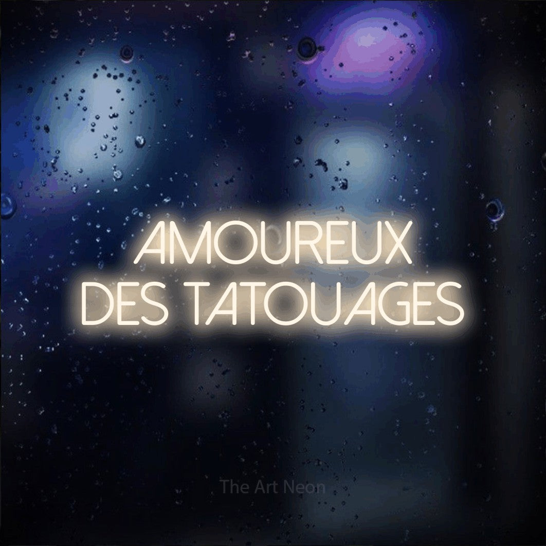“Amoureux des tatouages” LED neon sign in warm white, with thin sans-serif lettering across 2 lines, shown against glass with blue and purple reflections.