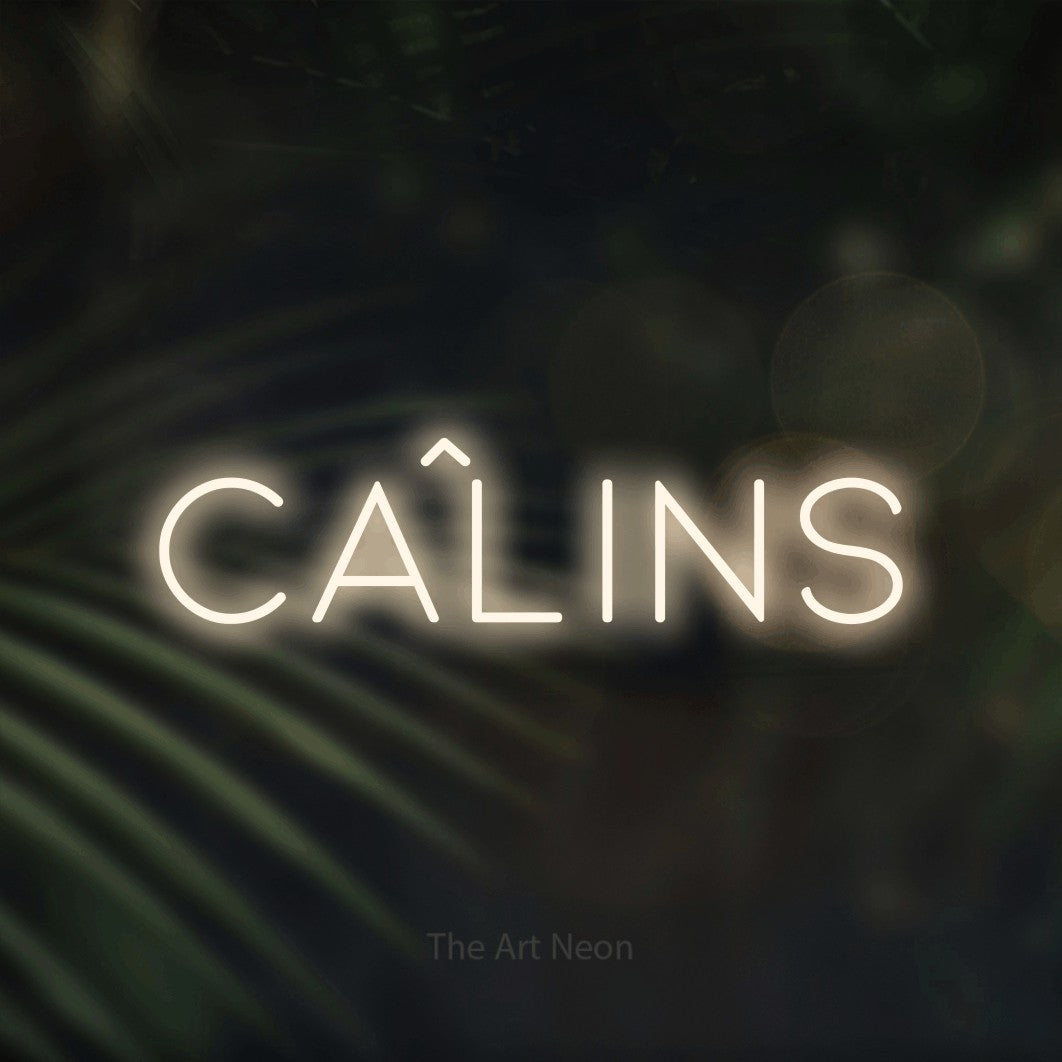 “Câlins” LED neon sign in warm white, with thin, widely spaced lettering, shown against a blurred palm and bokeh-light backdrop.